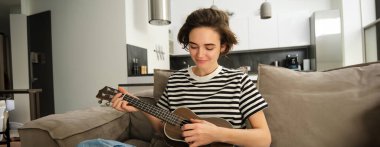 Genç modern kadın portresi, evde gitar çalan öğrenci, küçük bir gitarla oturan, şarkı söyleyen ve mutlu hisseden, kanepede oturan. Yaşam tarzı ve müzik konsepti