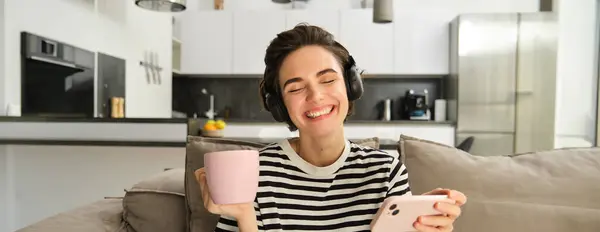 Carefree female model in headphones, sitting on sofa with smartphone and cup of tea, watching videos in wireless earphones on mobile phone app, resting at home.