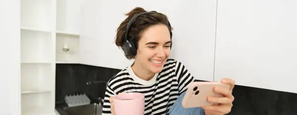 Portrait of woman watching funny videos on social media app, looking at smartphone and laughing, wearing headphones, drinking tea with cup, sitting on kitchen counter.