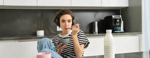 Cute girl eats her breakfast, holding bowl of cereals with milk, drinks coffee and listens to music in wireless headphones, using earphones while having her morning meal.