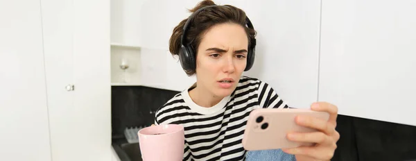 Portrait of woman with confused face, wearing headphones, watching tv series or show on smartphone app, frowning with clueless expression, drinking tea in the kitchen.