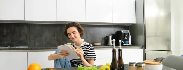 Portrait of young woman cooking, writing notes, grocery list in notebook, creating list of meals to cook through meal, sitting in kitchen near vegetables and chopping board.