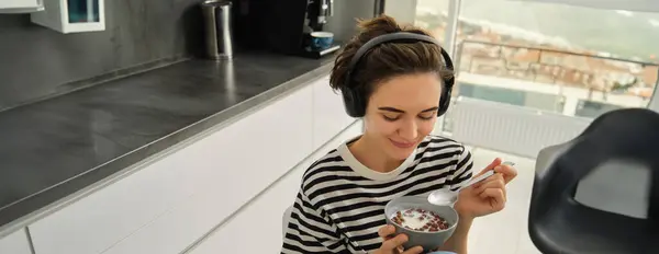 Close up portrait of smiling, beautiful young woman in headphones, eating cereals for breakfast and listening music or e-book, holding spoon with bowl.