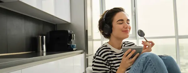 Portrait of smiling woman closes eyes from pleasure, listens music in headphones, eats breakfast, cereals with milk, holds spoon and bowl, sits in kitchen.
