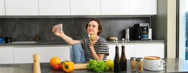 Mutfakta gülümseyen güzel bir kadının portresi, elmayla selfie çekmek, sebzelerle sağlıklı yemek pişirmek, yemek malzemelerini doğramak ve cep telefonuyla video kaydetmek..