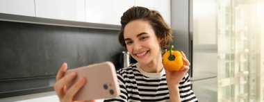 Gülümseyen, güzel genç bir kadın taze sarı biberle selfie çekiyor, yemek blogcusu mutfakta sebzelerle selfie çekiyor, vegan sosyal medya için yemek yapıyor ve video kaydediyor..
