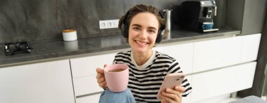 Çay içen ve kulaklıkla müzik dinleyen genç bir kadının portresi, mutfakta yemek yerken sosyal medyayı kaydırmak, mutlu bir şekilde gülümsemek..