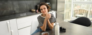 Gülümseyen modern kadın portresi, kahvaltıda süt ile mısır gevreği yemek, kablosuz kulaklıkla müzik dinlemek, evdeki mutfakta oturmak..