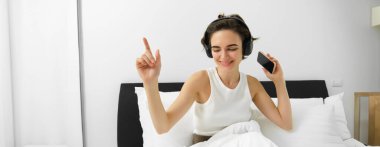 Yatağında dans eden, kablosuz kulaklıkla müzik dinleyen, cep telefonu tutan mutlu gülümseyen kaygısız bir kadın. Boşluğu kopyala