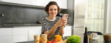 Genç bir kadının portresi mutfakta elinde sebzeler ve doğrama tahtasıyla duruyor, elinde akıllı telefon, cep telefonu tarayıcısında sağlıklı beslenme tarifleri arıyor, evde yemek pişiriyor..