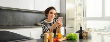 Güzel modern kadın portresi mutfakta yemek pişiriyor, akıllı telefon kullanıyor, yemek tarifi okuyor, kahvaltı için yemek arıyor, tezgaha sebzelerle yaslanıyor ve doğruyor..