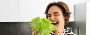 Mutlu, gülümseyen, salata yaprağı yiyen, sebzeleri seven, kendine salata yapan, mutfakta durup vejetaryen yemeği pişiren genç bir kadının yakın portresi..