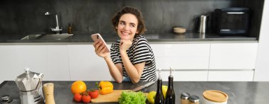 Sevimli genç esmer kadın mutfakta yemek pişiriyor, sebzeli sağlıklı yemek arıyor, akıllı telefon kullanıyor, tezgahın yanında oturuyor ve gülümsüyor..