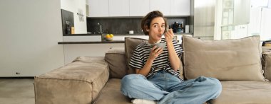 Kanepede şık genç bir kadının portresi, mısır gevreği yemek, kase ve kaşık tutmak, televizyon izlemek ve hızlı bir kahvaltı yapmak, evde oturma odasında vakit geçirmek..