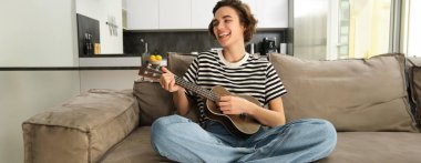 Ukuleleli neşeli genç bir kadın, müzik aleti çalıyor, küçük bir gitar tutuyor ve şarkı söylüyor, bacak bacak bacak üstüne atmış koltukta oturuyor, oturma odasında dinleniyor..