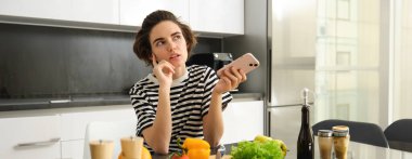 Genç kadın yemek blogcusu portresi, sosyal medya tarifinde paylaşım, vegan salatası, mutfakta akıllı telefonla durmak, sebze doğramak..