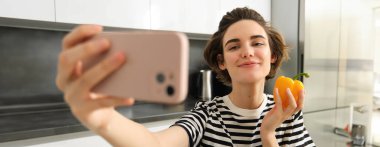 Gülümseyen, güzel genç bir kadın taze sarı biberle selfie çekiyor, yemek blogcusu mutfakta sebzelerle selfie çekiyor, vegan sosyal medya için yemek yapıyor ve video kaydediyor..
