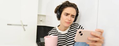 Hayal kırıklığına uğramış, isteksiz yüz ifadesi olan, akıllı telefondan sıkıcı bir dizi ya da film izleyen, kulaklık takan, çay içen ve mutfakta dinlenen bir kadın görüntüsü..