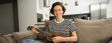 Tutkulu genç bir kadın, müzisyen evde ukulele çalıyor, neşeyle şarkı söylüyor, oturma odasında kanepede oturuyor..