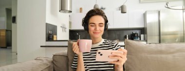 Genç bir kadın akıllı telefon uygulamasında videolar izliyor, kulaklıkla çay içiyor, kanepede oturuyor, evinde rahat vakit geçiriyor..