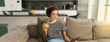 Kadın portresi evde oturuyor, dijital tabletle kablosuz kulaklık takıyor, taze portakal suyu içiyor..