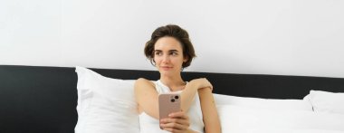 Gülümseyen esmer kadının portresi yatak odasında dinleniyor, cep telefonu kullanıyor ve evde kıyafetleriyle yatakta uzanıyor, mesajlaşıyor, akıllı telefon uygulamasından mesaj yolluyor..