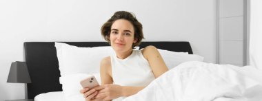 Genç bir kadının portresi yatakta uyanır, akıllı telefon kullanır, cep telefonunun alarmını kapatır, yatak odasında beyaz çarşafların üzerinde yatar..