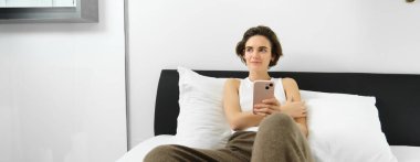 Yaşam tarzı ve teknoloji konsepti. Gülümseyen güzel bir kadın akıllı telefonuyla yatakta uzanıyor, yatak odasında dinleniyor, sosyal medyada sohbet ediyor, online alışveriş yapıyor..