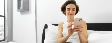 Gülümseyen güzel bir kadının portresi, dizüstü bilgisayarla yatakta oturmak, akıllı telefon kullanmak, cep telefonundan mesajları kontrol etmek, evde dinlenmek..