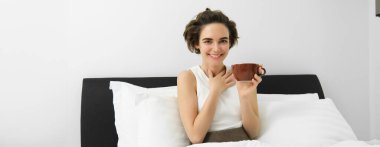 Bir fincan kahveyle, yatakta cappuccino içen ve gülümseyen güzel genç bir kadın..