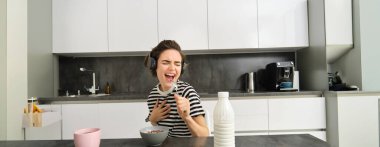 Mutlu ve duygusal genç kadın, kahvaltı yaparken şarkı söylüyor, mısır gevreğini sütle yiyor, kablosuz kulaklıkla müzik dinliyor, mutfakta oturuyor..