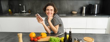 Mutluluktan gülümseyen bir kadın, akıllı telefonuyla mutfakta oturuyor, salata pişiriyor, masaya doğruyor, zeytinyağı ve sebze koyuyor..