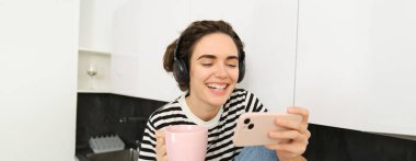 Yaşam tarzı konsepti. Genç modern kadın akıllı telefondan film izliyor, kulaklık takıyor, kahve içiyor ya da mutfakta çay içiyor..