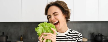 Mutlu ve sağlıklı genç bir kadının portresi, diyetini takip ediyor, marul yaprağı ile poz veriyor ve gülümsüyor, mutfakta yemek pişiriyor, vejetaryen sebzelerini seviyor..