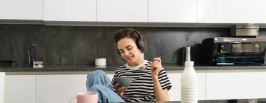 Mısır gevreğini sütle yiyen, çay içen ve kablosuz kulaklıkla müzik dinleyen mutlu bir kadının portresi, sabahının keyfini çıkarıyor, mutfakta tek başına kahvaltı yapıyor..