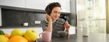 Güzel genç bir bayan model, mutfakta kahvaltı yapıyor, mısır gevreğini sütlü yiyor, gülümsüyor ve kahve içiyor, müzik dinliyor ya da kablosuz kulaklıkla podcast yapıyor..