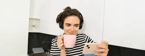 Smiling young woman watching tv series or video blog on smartphone app, drinking tea in the kitchen, looking at mobile phone with interest.