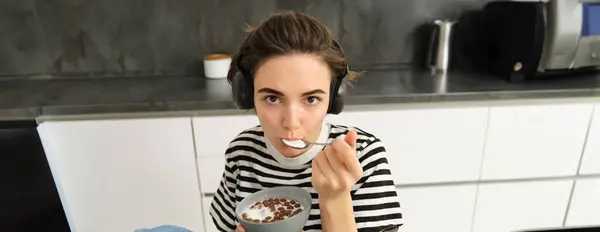 Beautiful woman, stylish girl eating cereals with milk, holding spoon near mouth and bowl in hands, having her breakfast in the morning, sitting in kitchen, listening music in headphones.