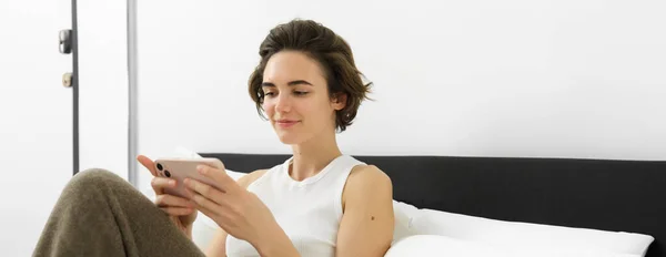 stock image Beautiful woman lying in bed with smartphone, resting in her hotel suite, using mobile phone for food delivery, reading on cellphone.