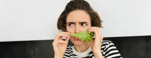 Portrait of woman frowning and grimacing, smelling fresh lettuce leaf with displeased face, standing in the kitchen, dislike vegetables.