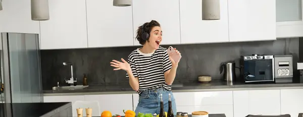 Portrait of woman singing and dancing while cooking food in the kitchen, holding smartphone, chopping vegetables on kitchen counter.