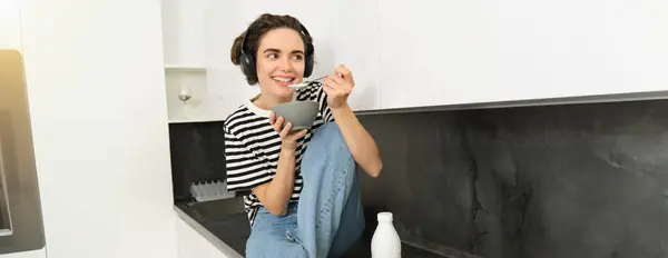 Close up portrait of young woman enjoys eating her breakfast, having cereals with milk, holding spoon and bowl, sits on kitchen counter with headphones, listens to music.
