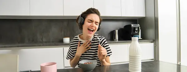 Carefree young woman, student in headphones, eating cereals for breakfast, listening music, singing and using soon as a microphone, having fun while having her meal in the morning.