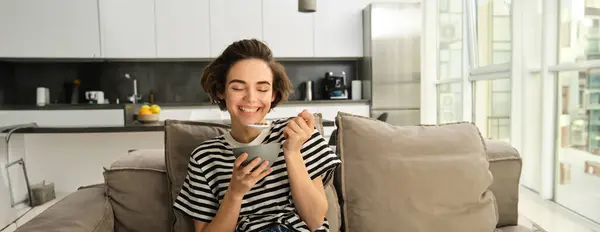 stock image Portrait of young modern woman, student eating cereals with milk and sitting on sofa, chilling in front of tv, having breakfast at home.