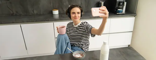 Portrait of modern happy woman, taking selfie, lifestyle blogger recording video of her eating breakfast and drinking tea in the kitchen, using smartphone for making content.
