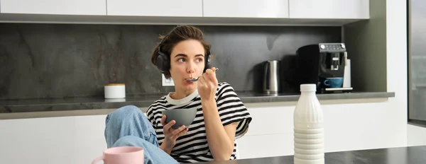 Cute modern woman, student eating quick breakfast, having cereals with milk and coffee, listening to podcast or music in wireless headphones.