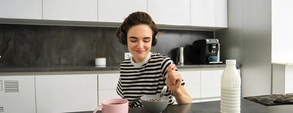 Cute modern woman, student eating quick breakfast, having cereals with milk and coffee, listening to podcast or music in wireless headphones.