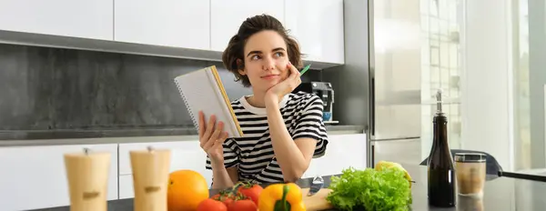 Portrait of thinking woman with notebook, cooking, writing down recipe ingredients, deciding on a meal for dinner, sitting near vegetables and chopping board in kitchen.