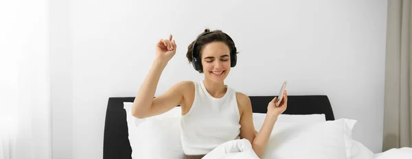 Portrait of happy young woman in bed, listening to music in wireless headphones, dancing in her bedroom, holding mobile phone in hand. Copy space