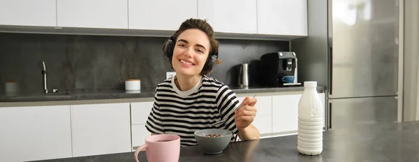 Portrait of happy smiling woman eating cereals with milk, drinking tea and listening music in wireless headphones, enjoys her morning, having breakfast alone in the kitchen.